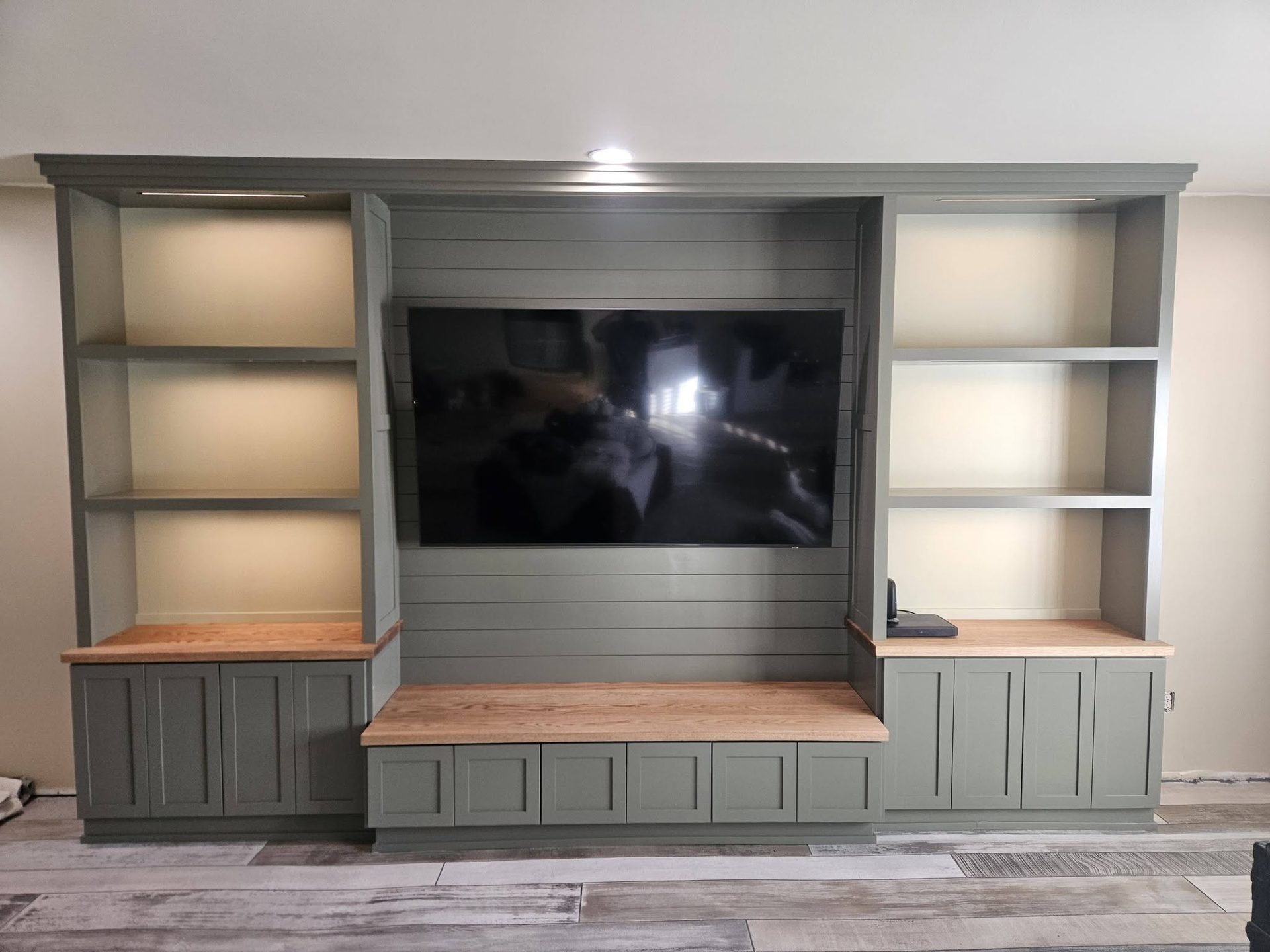 Built-in entertainment center with a wall-mounted TV on a shiplap panel, flanking open shelves lit from within, and a warm bench in solid wood like oak, maple, cherry, or walnut