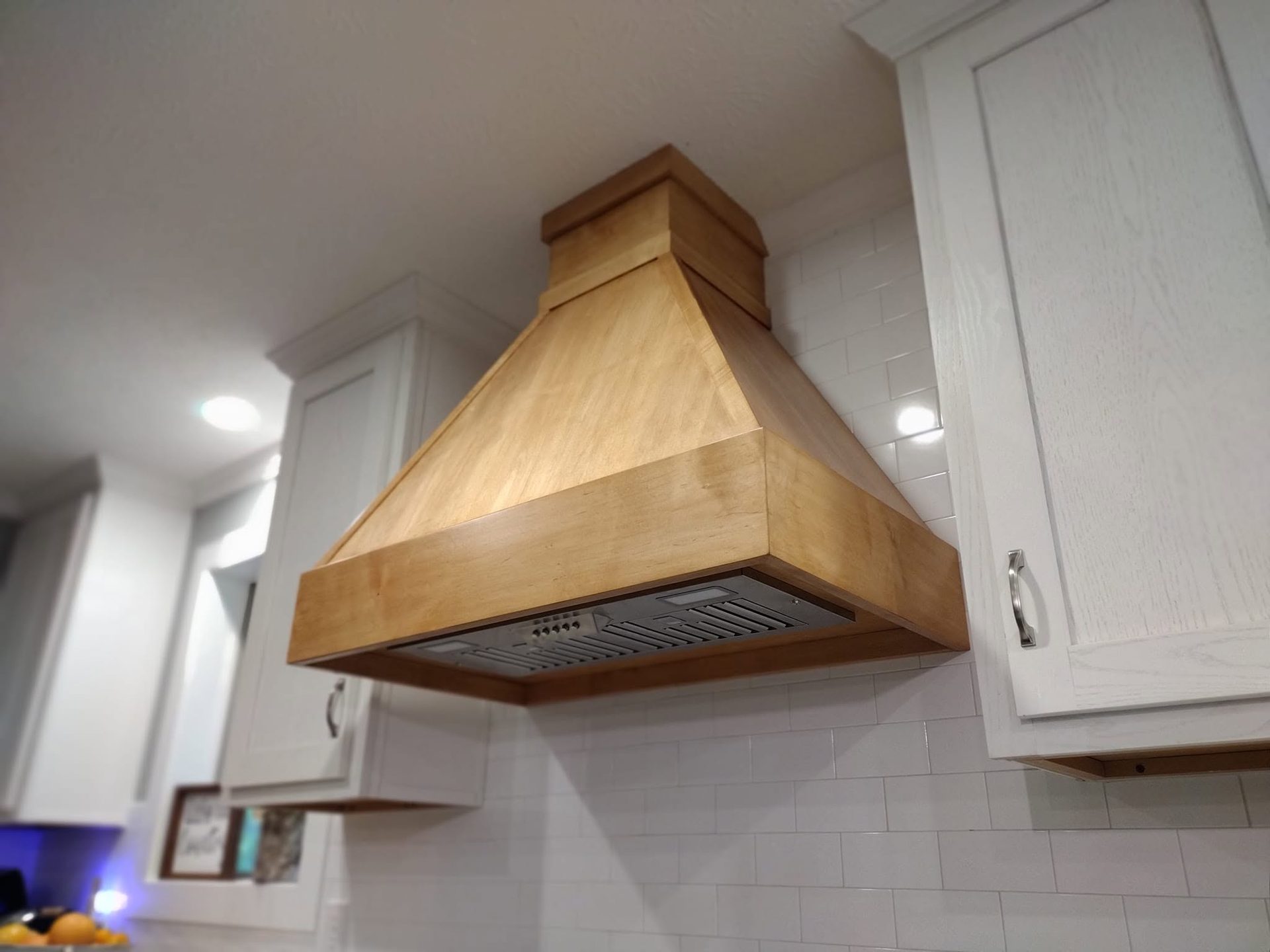 Close-up of a shop-built solid maple range hood mounted over white shaker cabinets and white subway tile