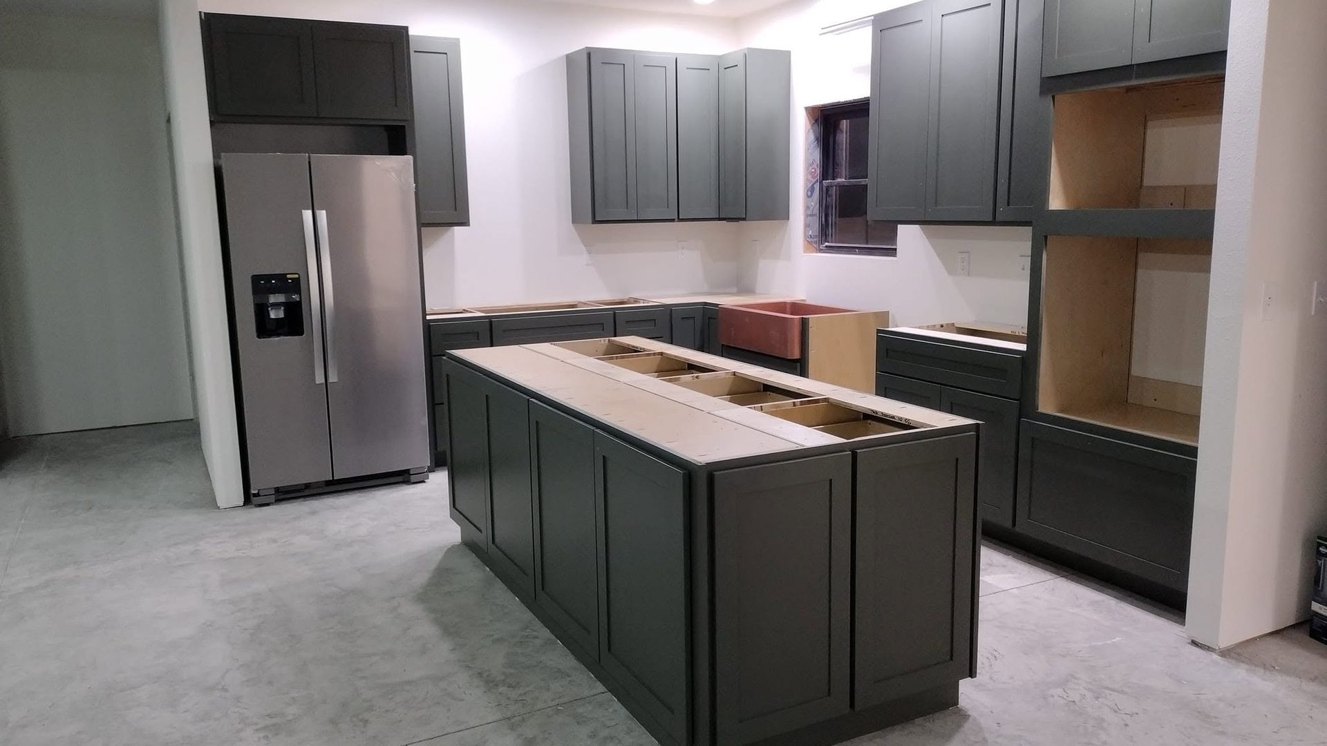 A kitchen mid-installation, with dark green-gray shaker cabinets, an island carcass being assembled, and a copper farmhouse apron sink set in place